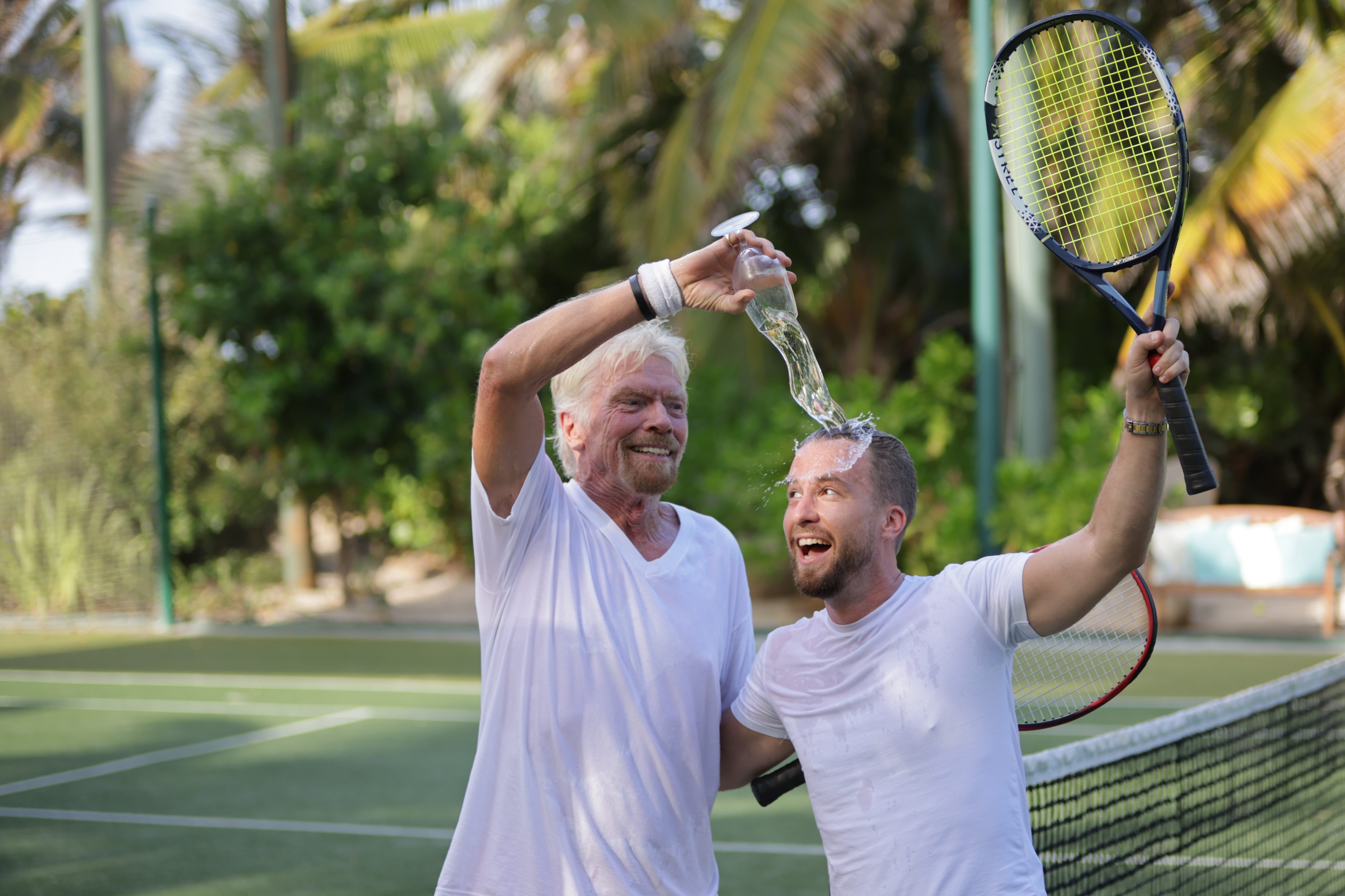 Richard Branson pouring water from a bottle over Jack Delosa with both holding tennis racquets on a tennis court