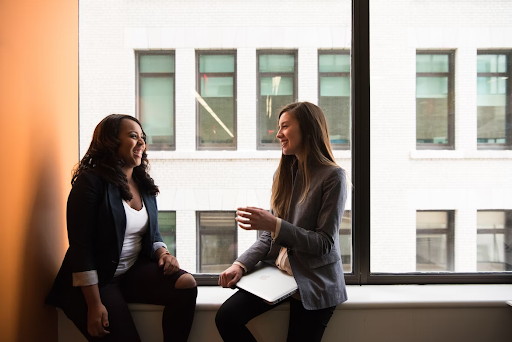 Two women sitting on a window ledge chatting